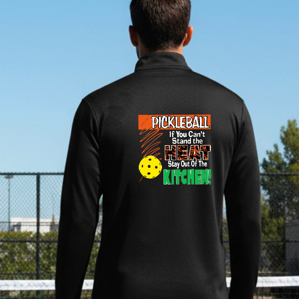 A man viewed from behind wearing a black long-sleeve quarter-zip pullover with a colorful pickleball-themed graphic on the back. The graphic features the word 'PICKLEBALL' in orange, a yellow pickleball, and the text 'If You Can't Stand the HEAT Stay Out Of The KITCHEN!' in white and green.
