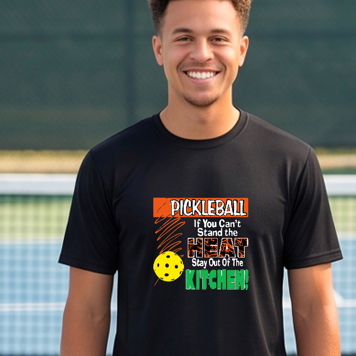 A man laughing on an outdoor pickleball court wearing a black t-shirt with a colorful graphic that reads 'PICKLEBALL If You Can't Stand the HEAT Stay Out Of The KITCHEN!'.