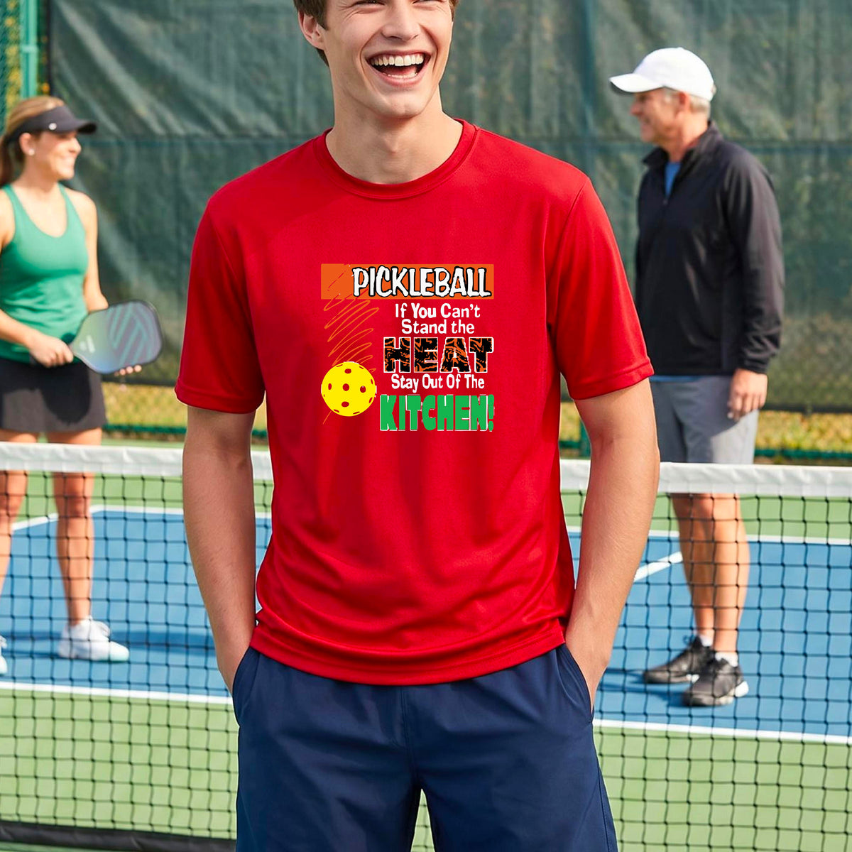 A man laughing on an outdoor pickleball court wearing a red t-shirt with a colorful graphic that reads 'PICKLEBALL If You Can't Stand the HEAT Stay Out Of The KITCHEN!'.