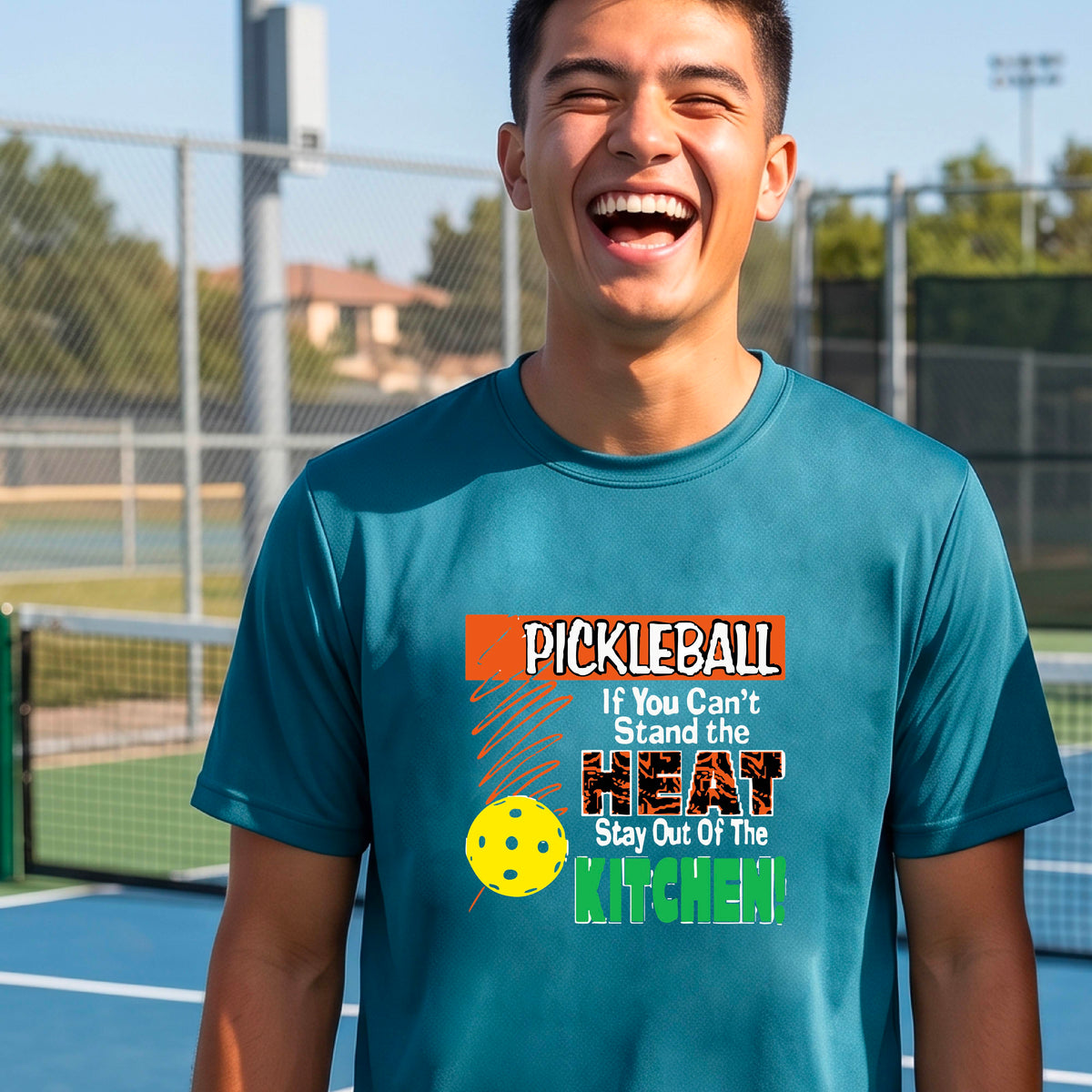 A man laughing on an outdoor pickleball court wearing a teal t-shirt with a colorful graphic that reads 'PICKLEBALL If You Can't Stand the HEAT Stay Out Of The KITCHEN!'.