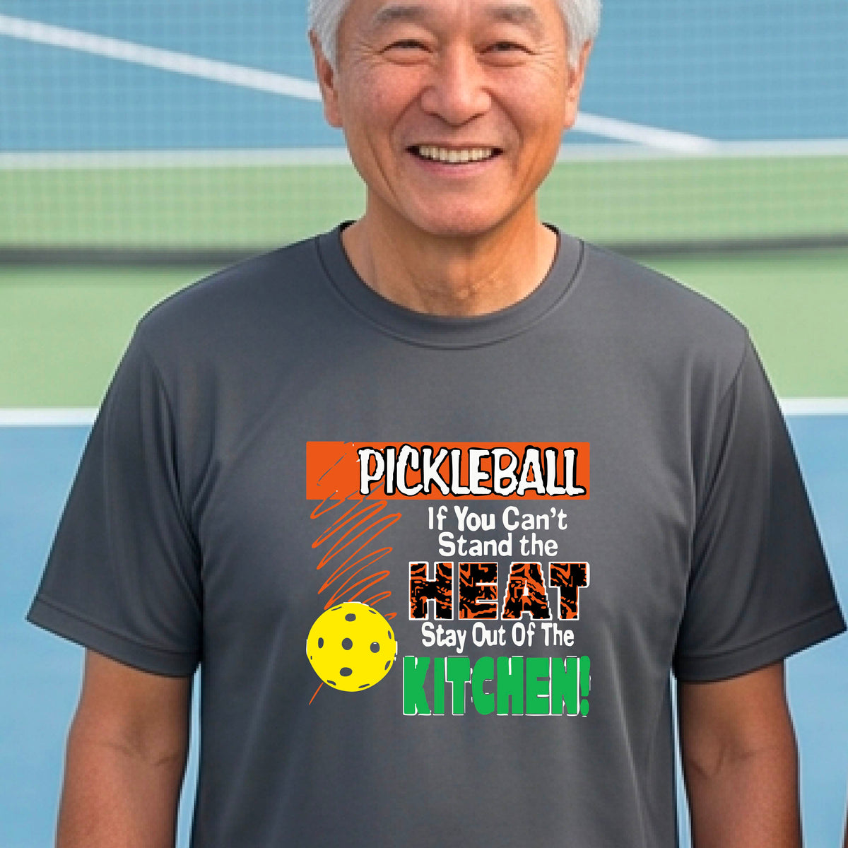 A man laughing on an outdoor pickleball court wearing a gray t-shirt with a colorful graphic that reads 'PICKLEBALL If You Can't Stand the HEAT Stay Out Of The KITCHEN!'.