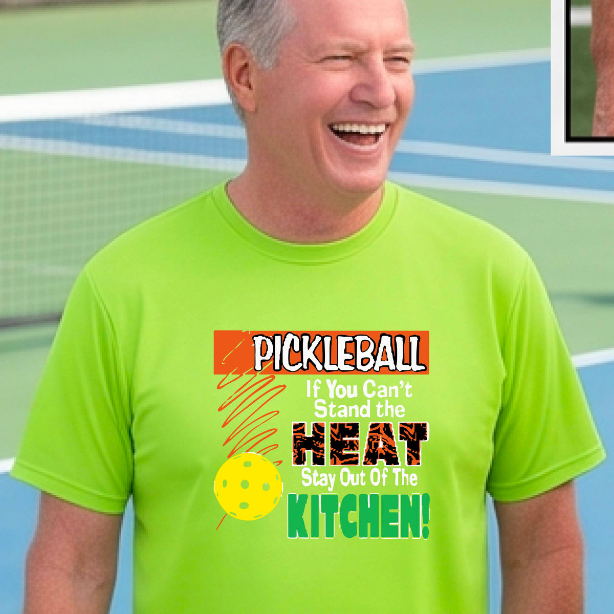 A man laughing on an outdoor pickleball court wearing a lime green t-shirt with a colorful graphic that reads 'PICKLEBALL If You Can't Stand the HEAT Stay Out Of The KITCHEN!'.
