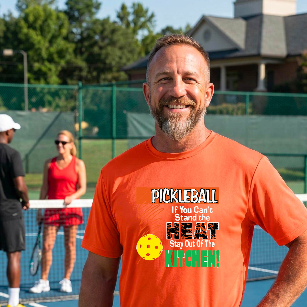 A man laughing on an outdoor pickleball court wearing an orange t-shirt with a colorful graphic that reads 'PICKLEBALL If You Can't Stand the HEAT Stay Out Of The KITCHEN!'.