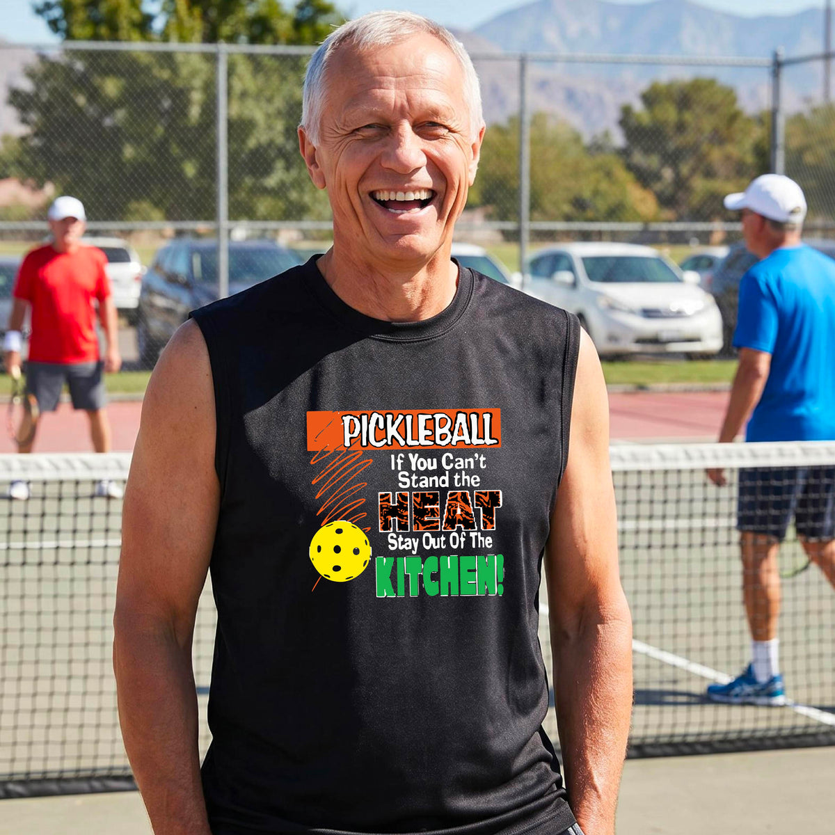 A man wearing a bright black sleeveless t-shirt with a colorful pickleball graphic and text on the front, “If You Can't Stand The Heat Stay Out Of The Kitchen" set against a blurred outdoor pickleball court background.