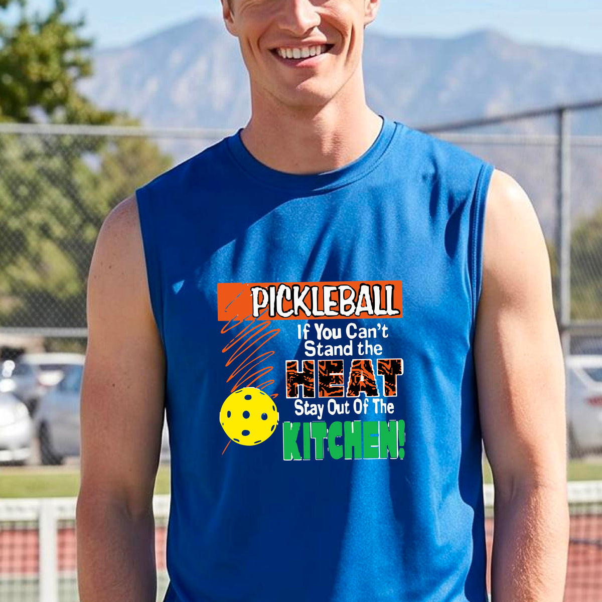 A man wearing a bright royal blue sleeveless t-shirt with a colorful pickleball graphic and text on the front, “If You Can't Stand The Heat Stay Out Of The Kitchen" set against a blurred outdoor pickleball court background.