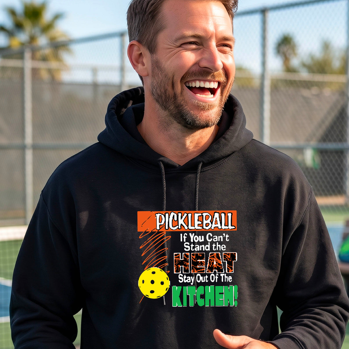 A man wearing a black hoodie with a colorful pickleball graphic on the chest with text “If You Can't Stand The Heat Stay Out Of The Kitchen", standing on a blurred blue pickleball court.