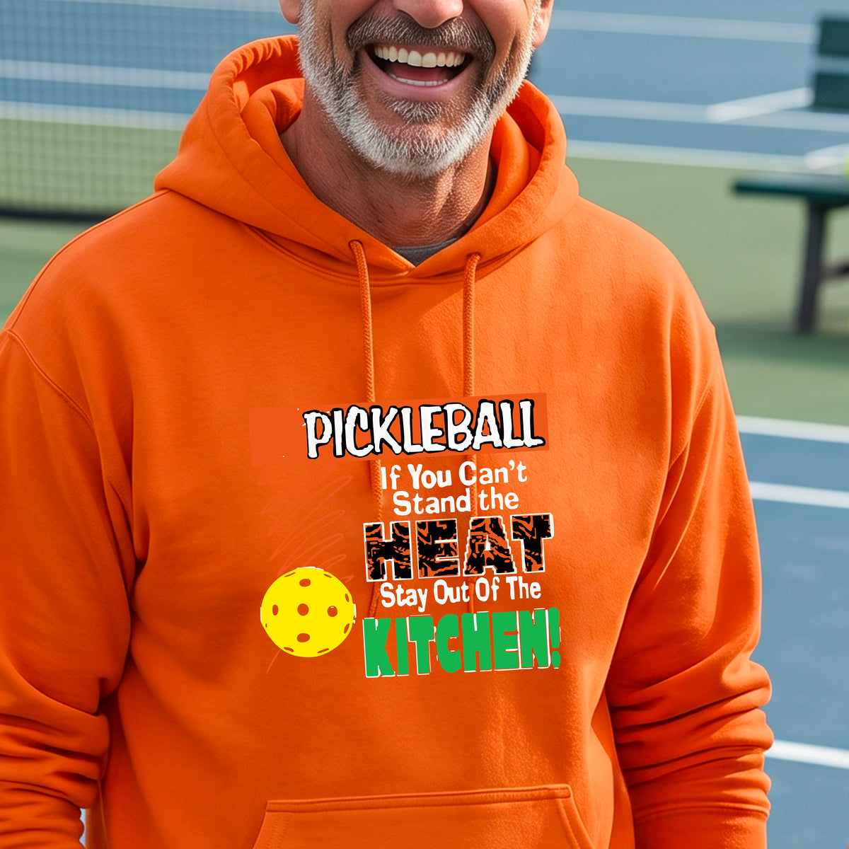 A man wearing a bright orange hoodie with a colorful pickleball graphic on the chest with text “If You Can't Stand The Heat Stay Out Of The Kitchen", standing on a blurred blue pickleball court.