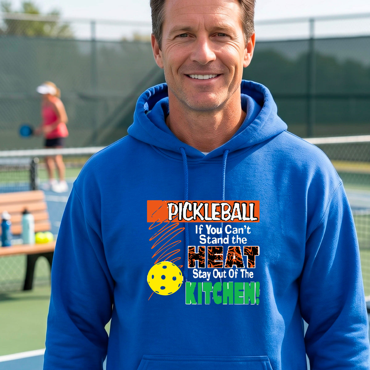 A man wearing a bright blue hoodie with a colorful pickleball graphic on the chest with text “If You Can't Stand The Heat Stay Out Of The Kitchen", standing on a blurred blue pickleball court.