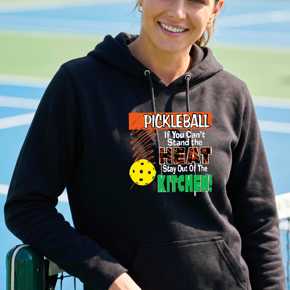A woman wearing a bright black hoodie with a colorful pickleball graphic on the chest with text “If You Can't Stand The Heat Stay Out Of The Kitchen", standing on a blurred blue pickleball court.