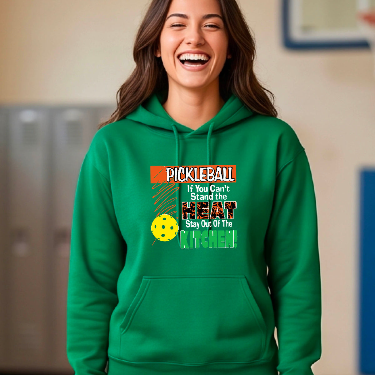 A woman wearing a bright green hoodie with a colorful pickleball graphic on the chest with text “If You Can't Stand The Heat Stay Out Of The Kitchen", standing on a blurred blue pickleball court.