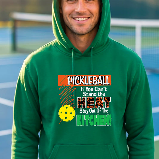 A man wearing a bright green hoodie with a colorful pickleball graphic on the chest with text “If You Can't Stand The Heat Stay Out Of The Kitchen", standing on a blurred blue pickleball court.