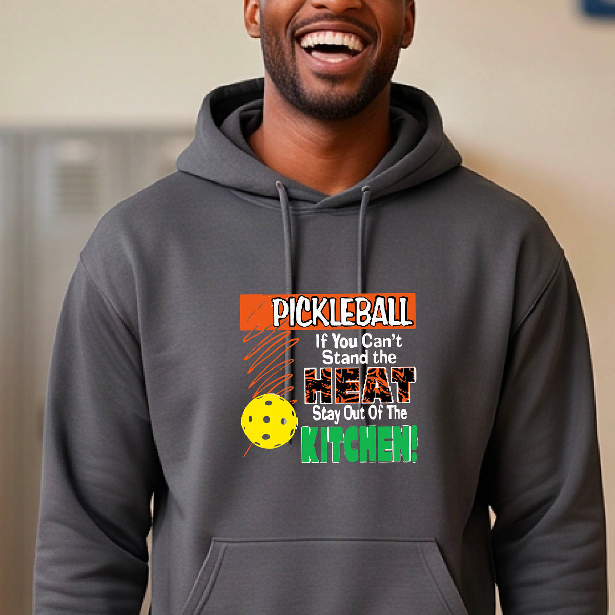 A man wearing a gray hoodie with a colorful pickleball graphic on the chest with text “If You Can't Stand The Heat Stay Out Of The Kitchen", standing on a blurred blue pickleball court.