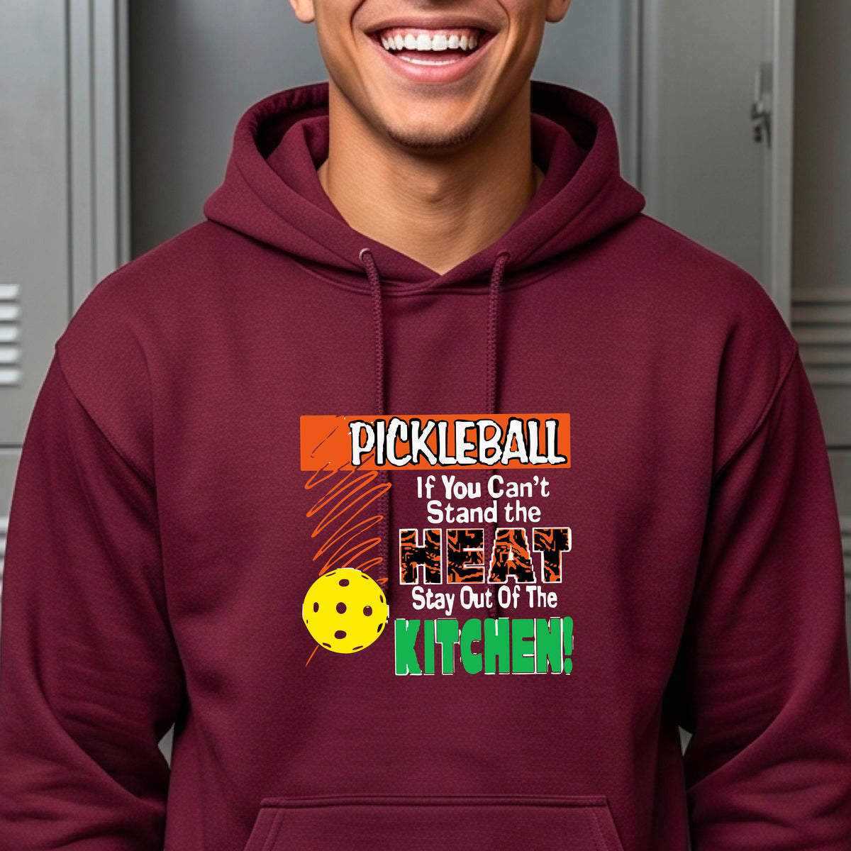 A man wearing a maroon hoodie with a colorful pickleball graphic on the chest with text “If You Can't Stand The Heat Stay Out Of The Kitchen", standing on a blurred blue pickleball court.