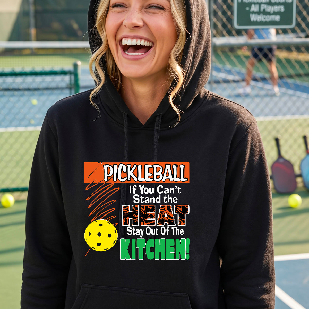 A woman wearing a black hoodie with a colorful pickleball graphic and text that reads 'PICKLEBALL If You Can't Stand the HEAT Stay Out Of The KITCHEN!' on a pickleball court background.