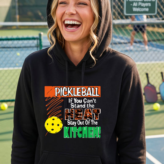A woman wearing a black hoodie with a colorful pickleball graphic and text that reads 'PICKLEBALL If You Can't Stand the HEAT Stay Out Of The KITCHEN!' on a pickleball court background.