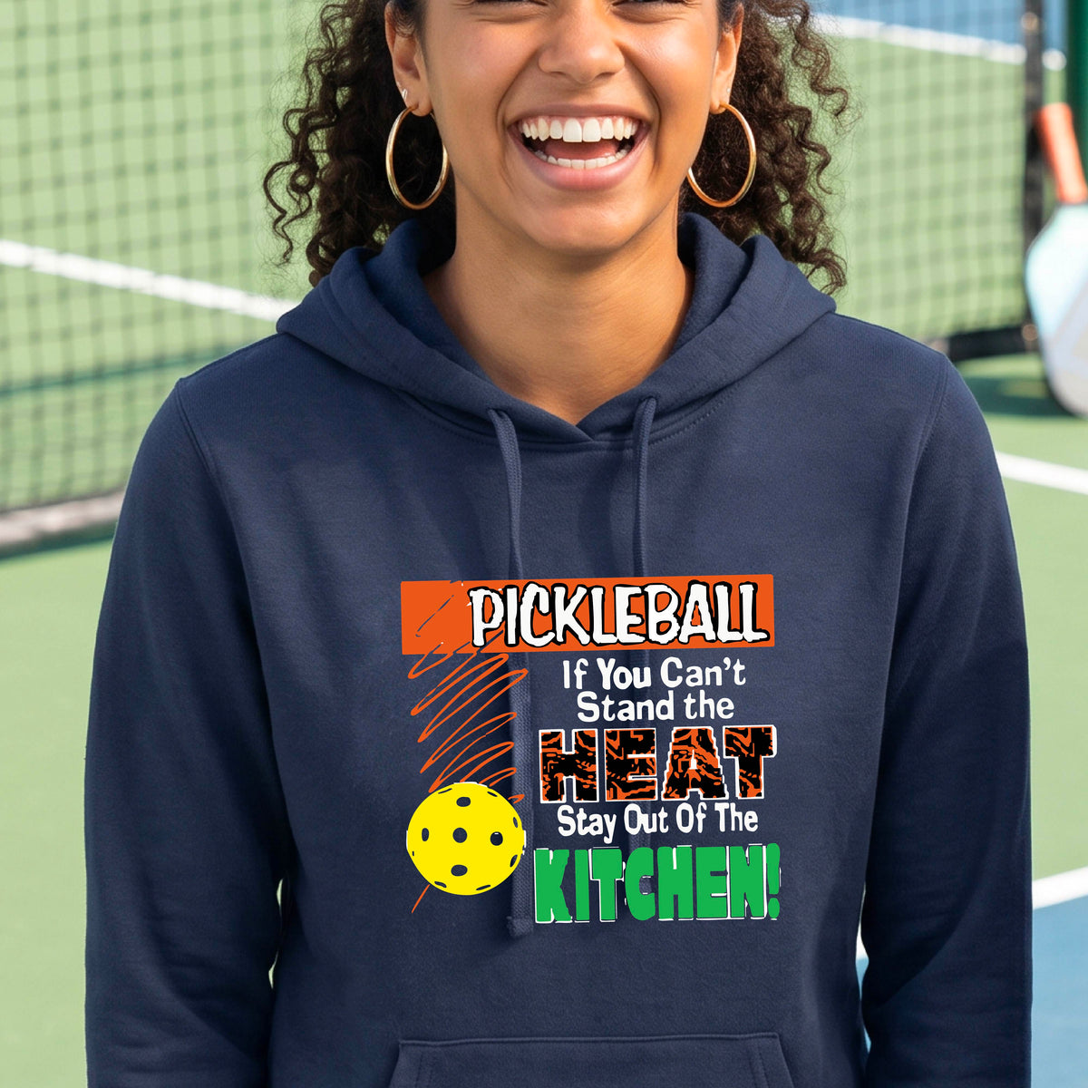 A woman wearing a navy blue hoodie with a colorful pickleball graphic and text that reads 'PICKLEBALL If You Can't Stand the HEAT Stay Out Of The KITCHEN!' on a pickleball court background.