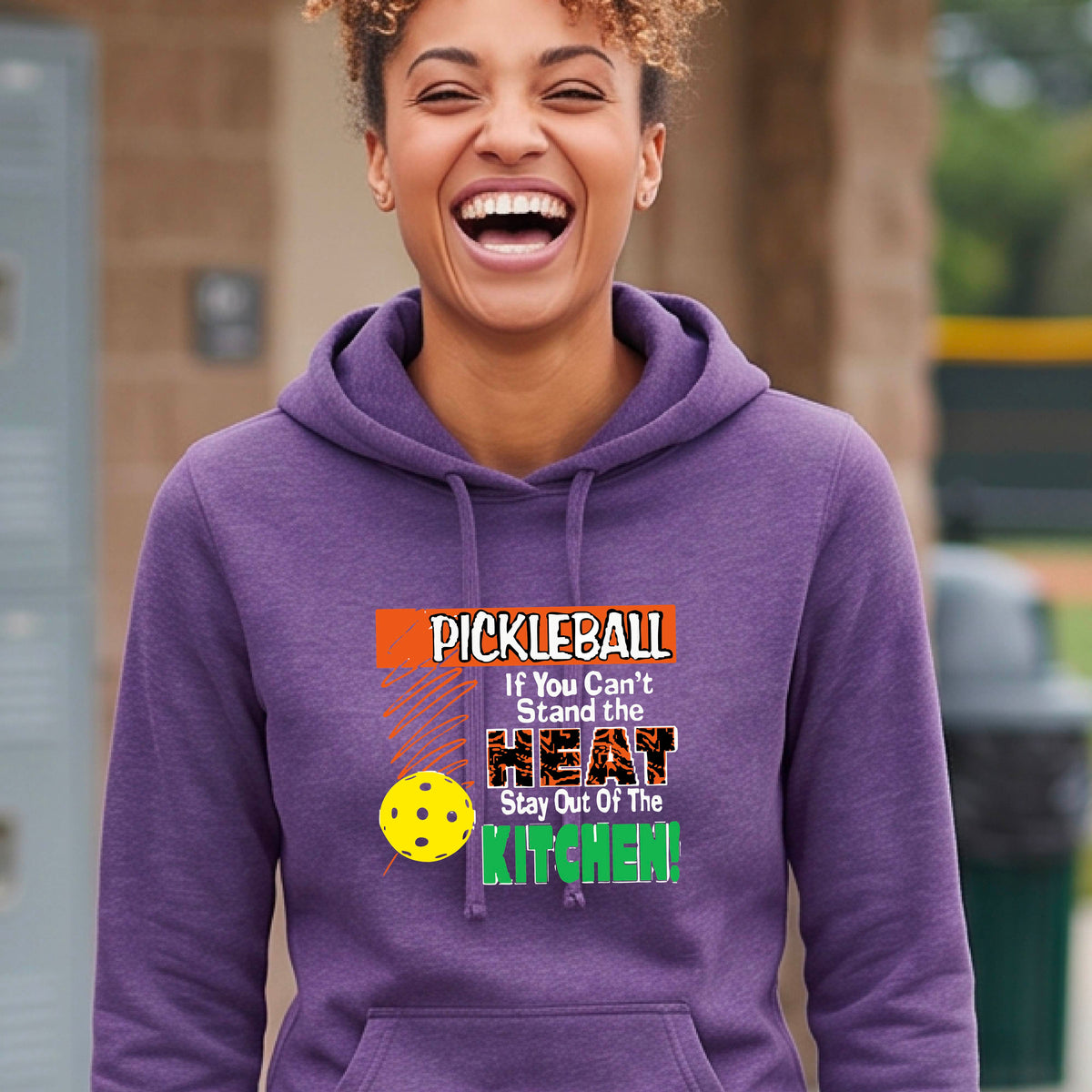 A woman wearing a purple hoodie with a colorful pickleball graphic and text that reads 'PICKLEBALL If You Can't Stand the HEAT Stay Out Of The KITCHEN!' on a pickleball court background.