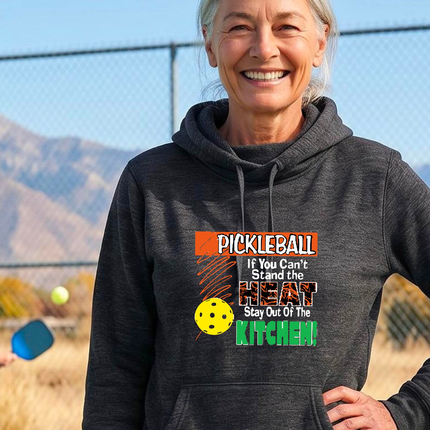 A smiling woman outdoors wearing a heather black cowl-neck hoodie with a colorful pickleball graphic and text on the front “If You Can't Stand The Heat Stay Out Of The Kitchen"