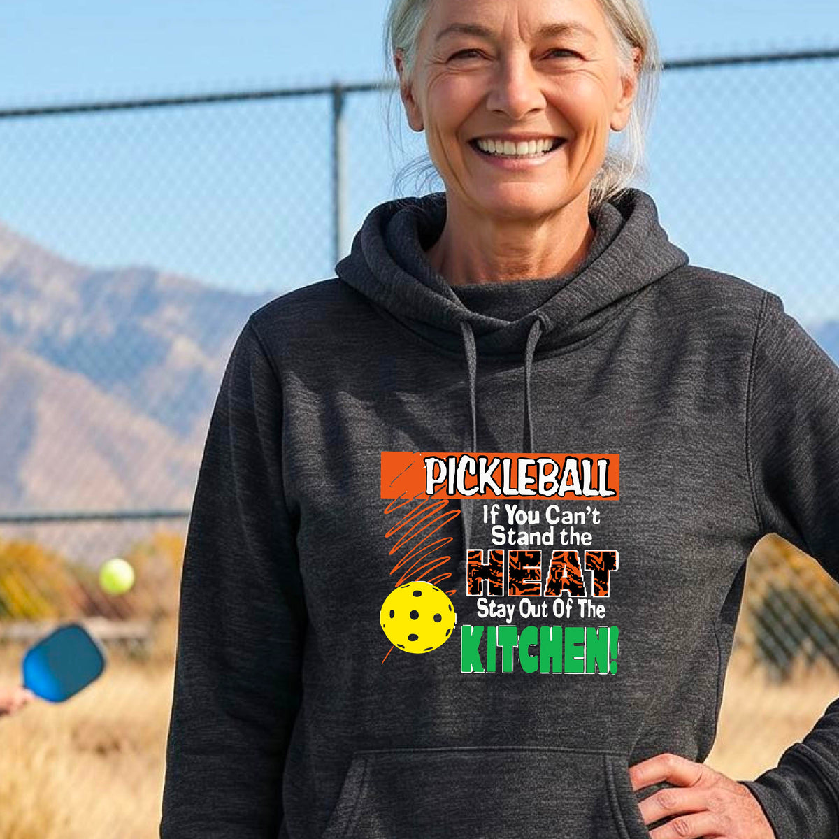 A smiling woman outdoors wearing a heather black cowl-neck hoodie with a colorful pickleball graphic and text on the front “If You Can't Stand The Heat Stay Out Of The Kitchen"
