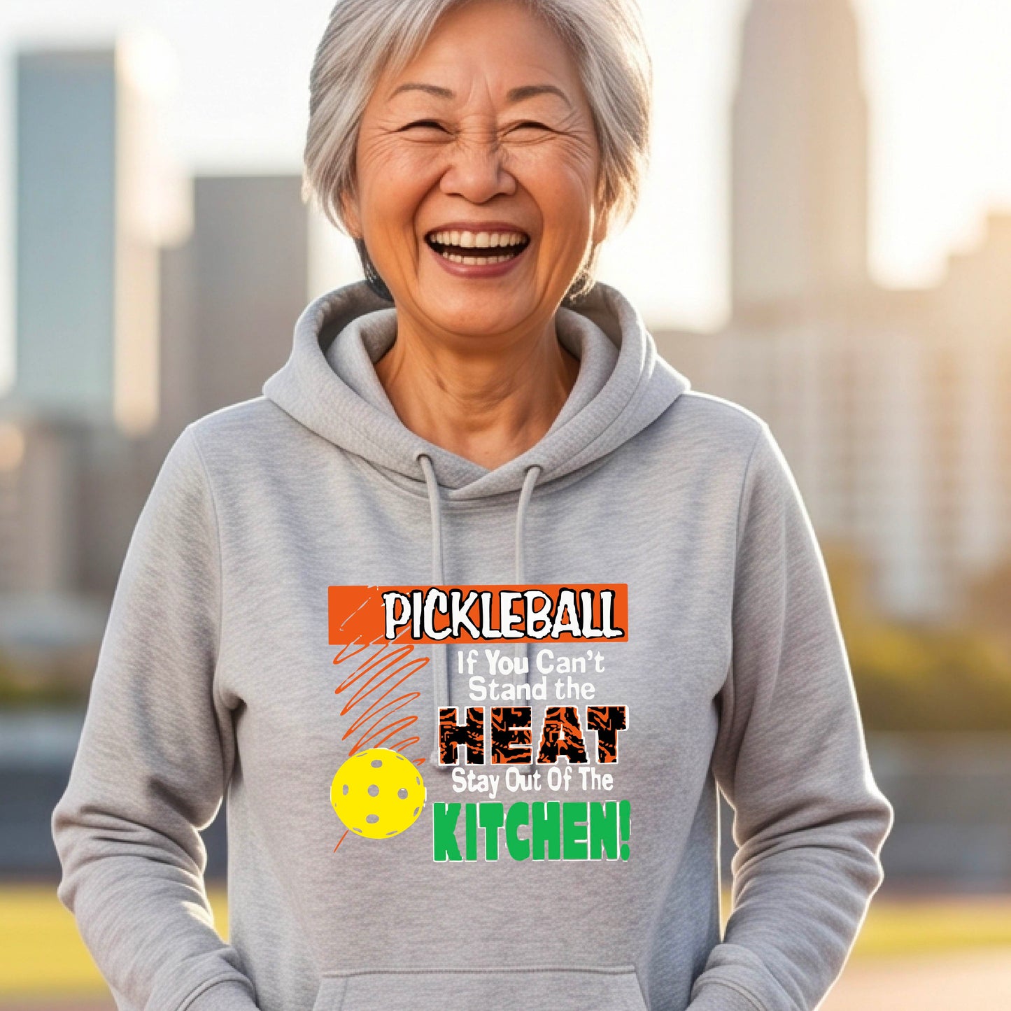 A smiling woman outdoors wearing a gray cowl-neck hoodie with a colorful pickleball graphic and text on the front “If You Can't Stand The Heat Stay Out Of The Kitchen"