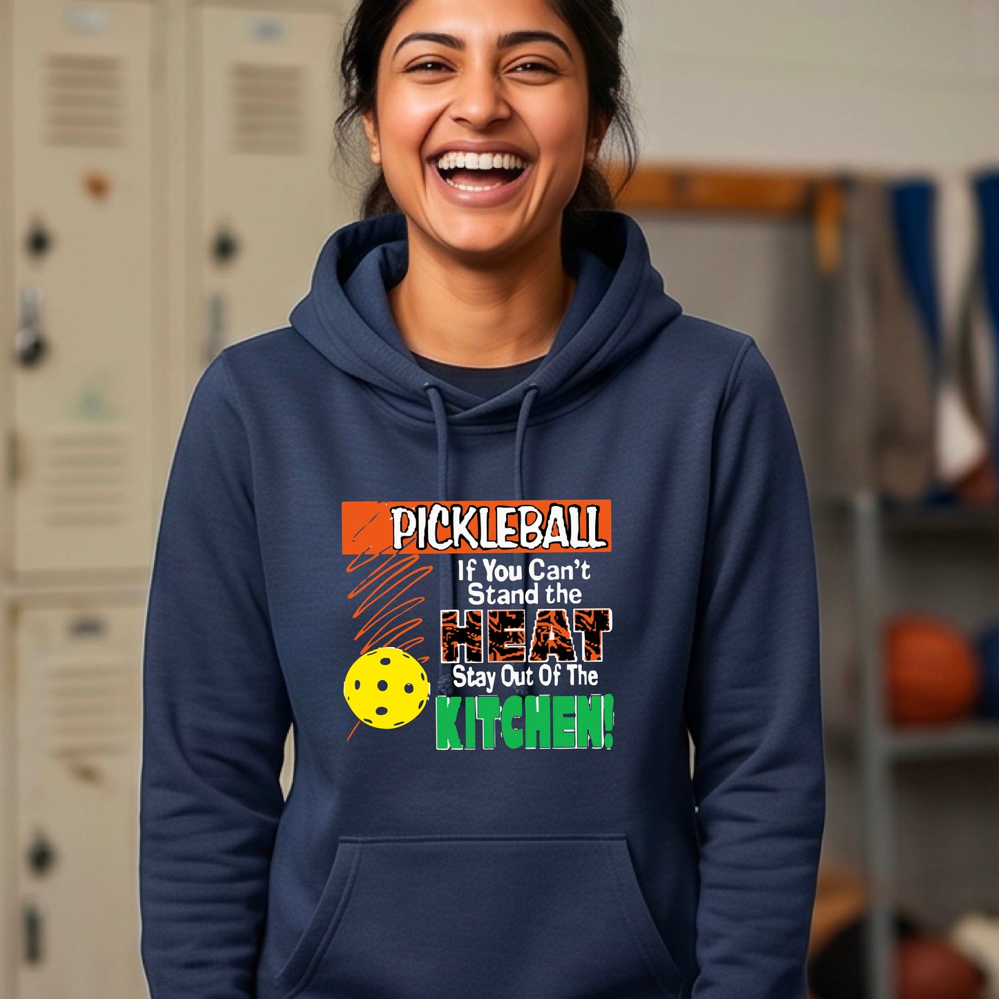 A smiling woman outdoors wearing a navy blue cowl-neck hoodie with a colorful pickleball graphic and text on the front “If You Can't Stand The Heat Stay Out Of The Kitchen"