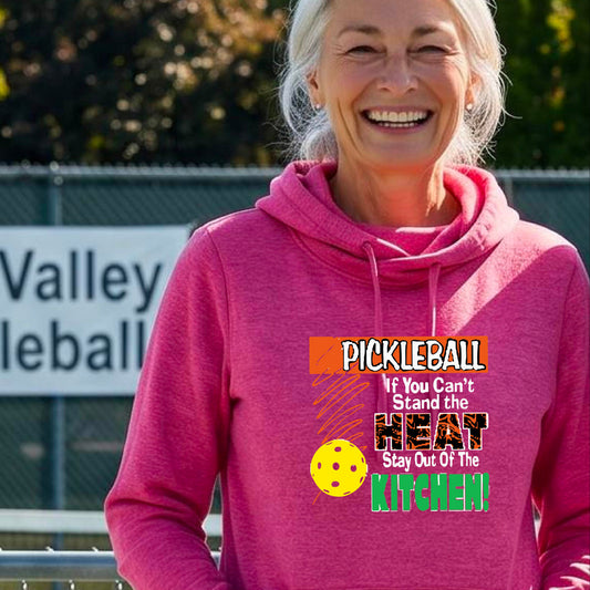 A smiling woman outdoors wearing a bright pink cowl-neck hoodie with a colorful pickleball graphic and text on the front “If You Can't Stand The Heat Stay Out Of The Kitchen"