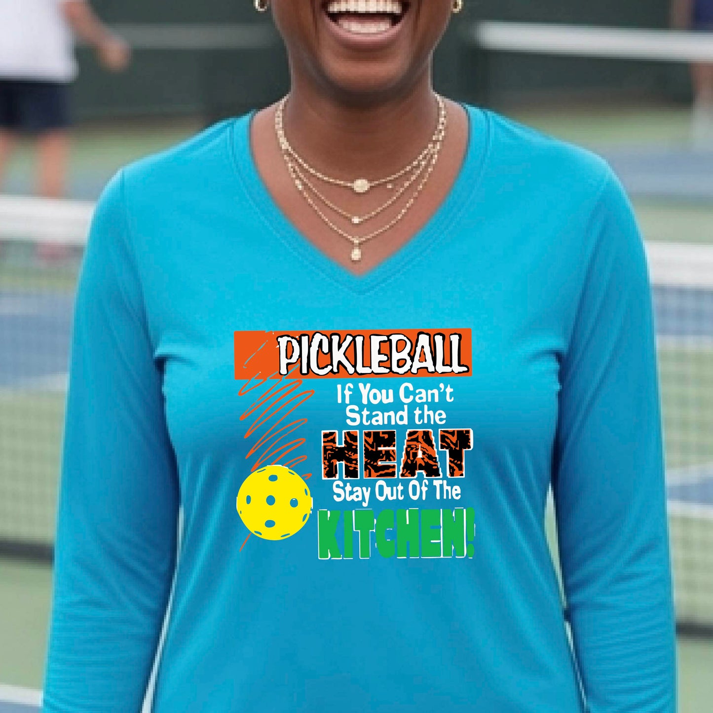 A woman wearing a light blue long-sleeve V-neck t-shirt with a colorful pickleball graphic and text on the front, "PICKLEALL IF YOU CAN'T STAND THE HEAT STAY OUT OF THE KITCHEN", standing on an outdoor pickleball court.