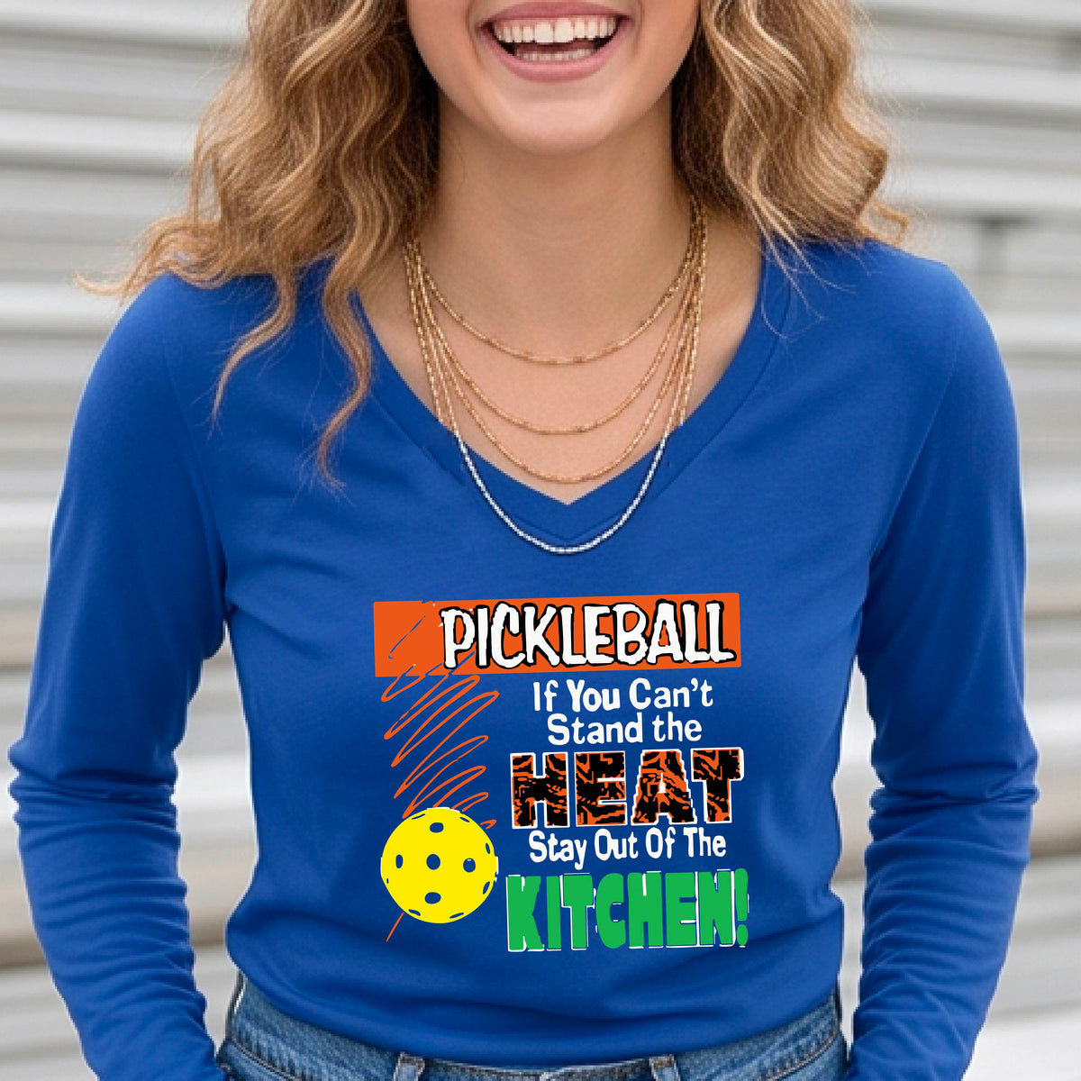 A woman wearing a royal blue long-sleeve V-neck t-shirt with a colorful pickleball graphic and text on the front, "PICKLEALL IF YOU CAN'T STAND THE HEAT STAY OUT OF THE KITCHEN", standing on an outdoor pickleball coURT.
