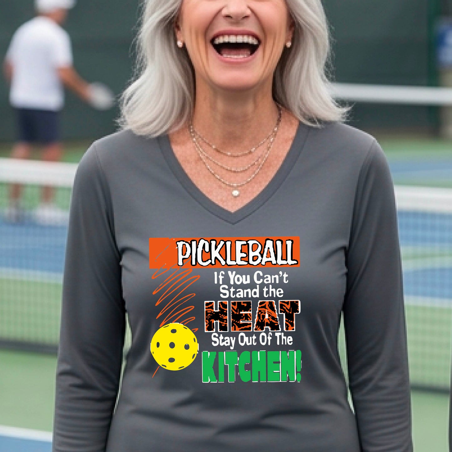 A woman wearing a gray long-sleeve V-neck t-shirt with a colorful pickleball graphic and text on the front, "PICKLEALL IF YOU CAN'T STAND THE HEAT STAY OUT OF THE KITCHEN", standing on an outdoor pickleball court.