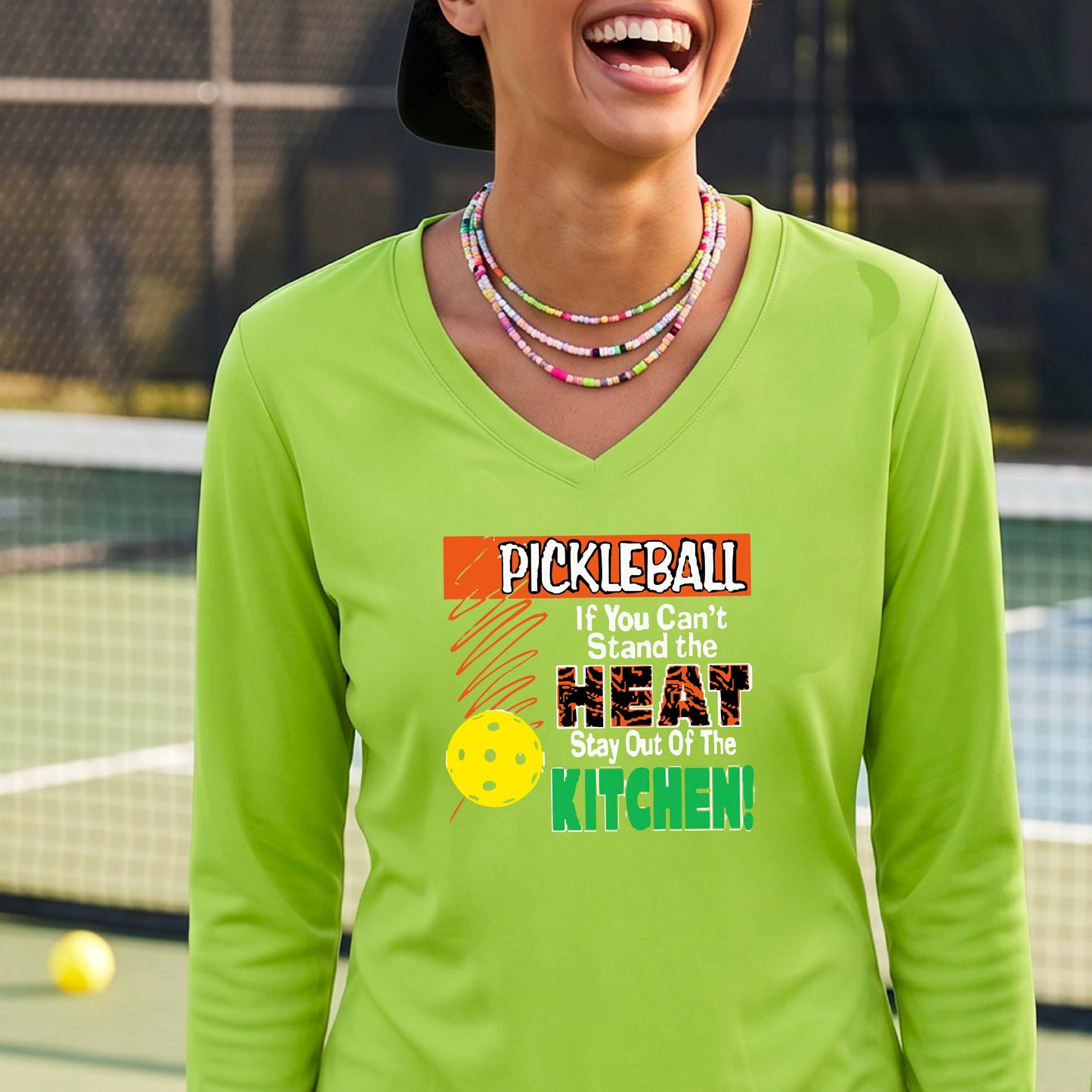 A woman wearing a lime green long-sleeve V-neck t-shirt with a colorful pickleball graphic and text on the front, "PICKLEALL IF YOU CAN'T STAND THE HEAT STAY OUT OF THE KITCHEN", standing on an outdoor pickleball court.