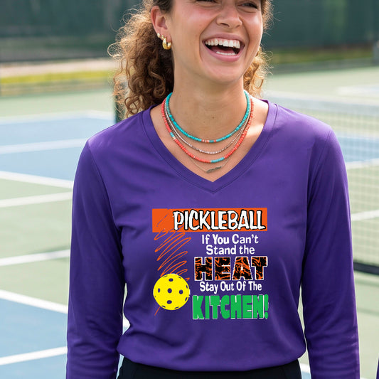 A woman wearing a purple long-sleeve V-neck t-shirt with a colorful pickleball graphic and text on the front, "PICKLEALL IF YOU CAN'T STAND THE HEAT STAY OUT OF THE KITCHEN", standing on an outdoor pickleball court.