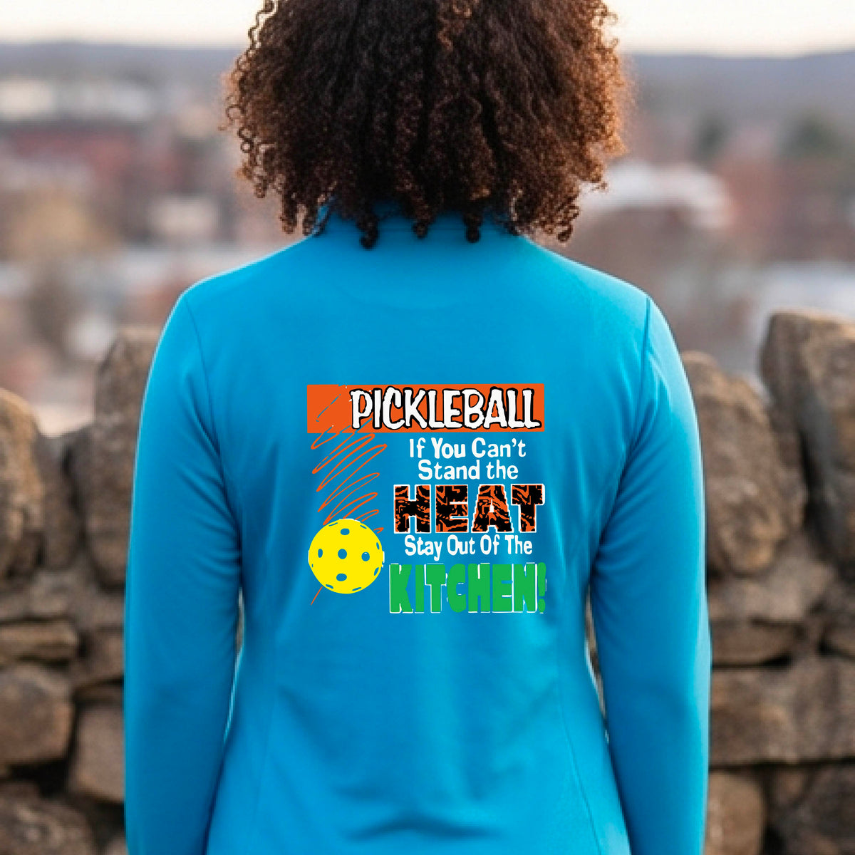 A person wearing a bright blue athletic quarter-zip jacket, shown from the back on a blue pickleball court. The back of the jacket features a colorful graphic with the text 'PICKLEBALL If You Can't Stand the HEAT Stay Out Of The KITCHEN!' and a yellow pickleball illustration.