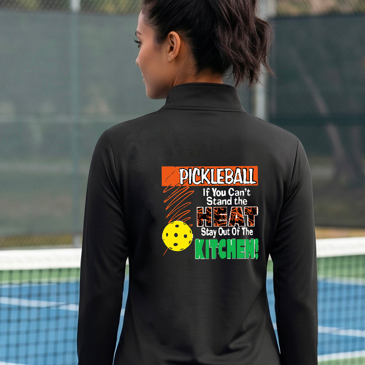 A person wearing a black athletic quarter-zip jacket, shown from the back on a blue pickleball court. The back of the jacket features a colorful graphic with the text 'PICKLEBALL If You Can't Stand the HEAT Stay Out Of The KITCHEN!' and a yellow pickleball illustration.