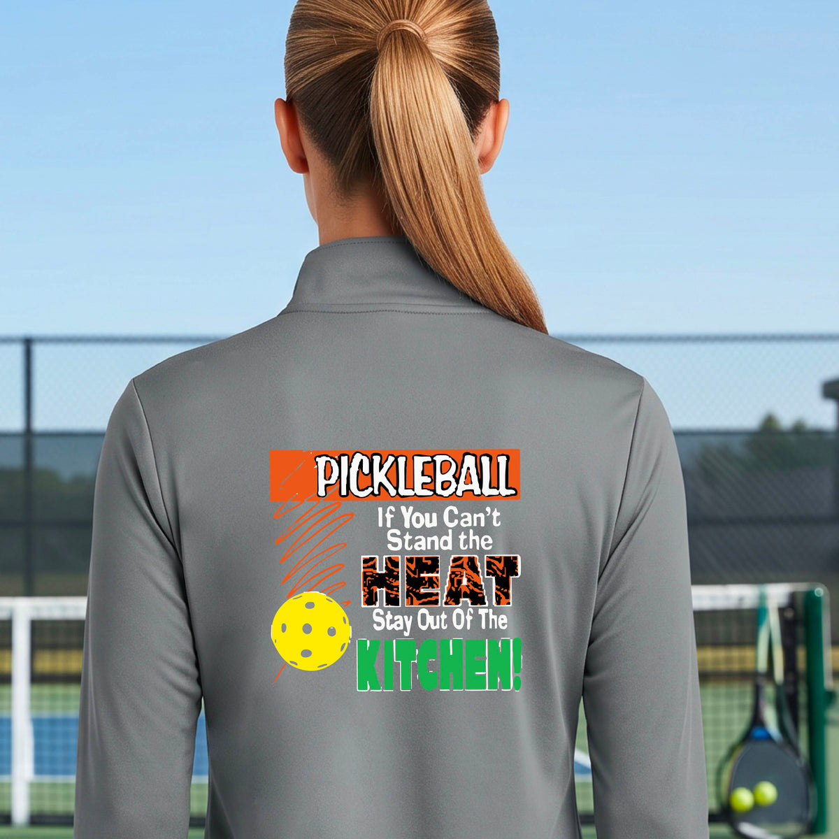 A person wearing a gray athletic quarter-zip jacket, shown from the back on a blue pickleball court. The back of the jacket features a colorful graphic with the text 'PICKLEBALL If You Can't Stand the HEAT Stay Out Of The KITCHEN!' and a yellow pickleball illustration.