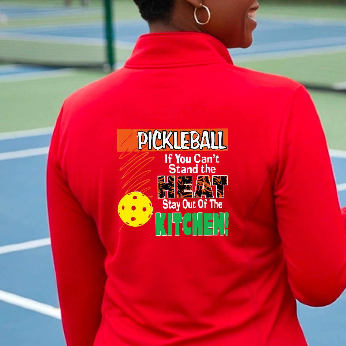 A person wearing a bright red athletic quarter-zip jacket, shown from the back on a blue pickleball court. The back of the jacket features a colorful graphic with the text 'PICKLEBALL If You Can't Stand the HEAT Stay Out Of The KITCHEN!' and a yellow pickleball illustration.