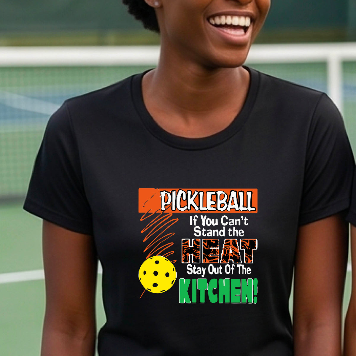 A woman wearing a black short-sleeve t-shirt with a colorful graphic that reads 'PICKLEBALL If You Can't Stand the HEAT Stay Out Of The KITCHEN!' on a pickleball court background.