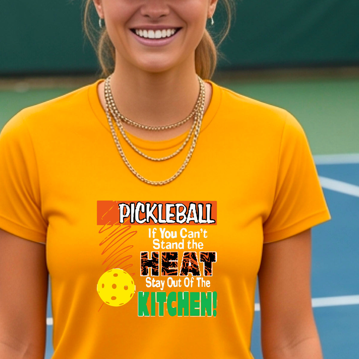 A woman wearing a mustard yellow gold short-sleeve t-shirt with a colorful graphic that reads 'PICKLEBALL If You Can't Stand the HEAT Stay Out Of The KITCHEN!' on a pickleball court background.