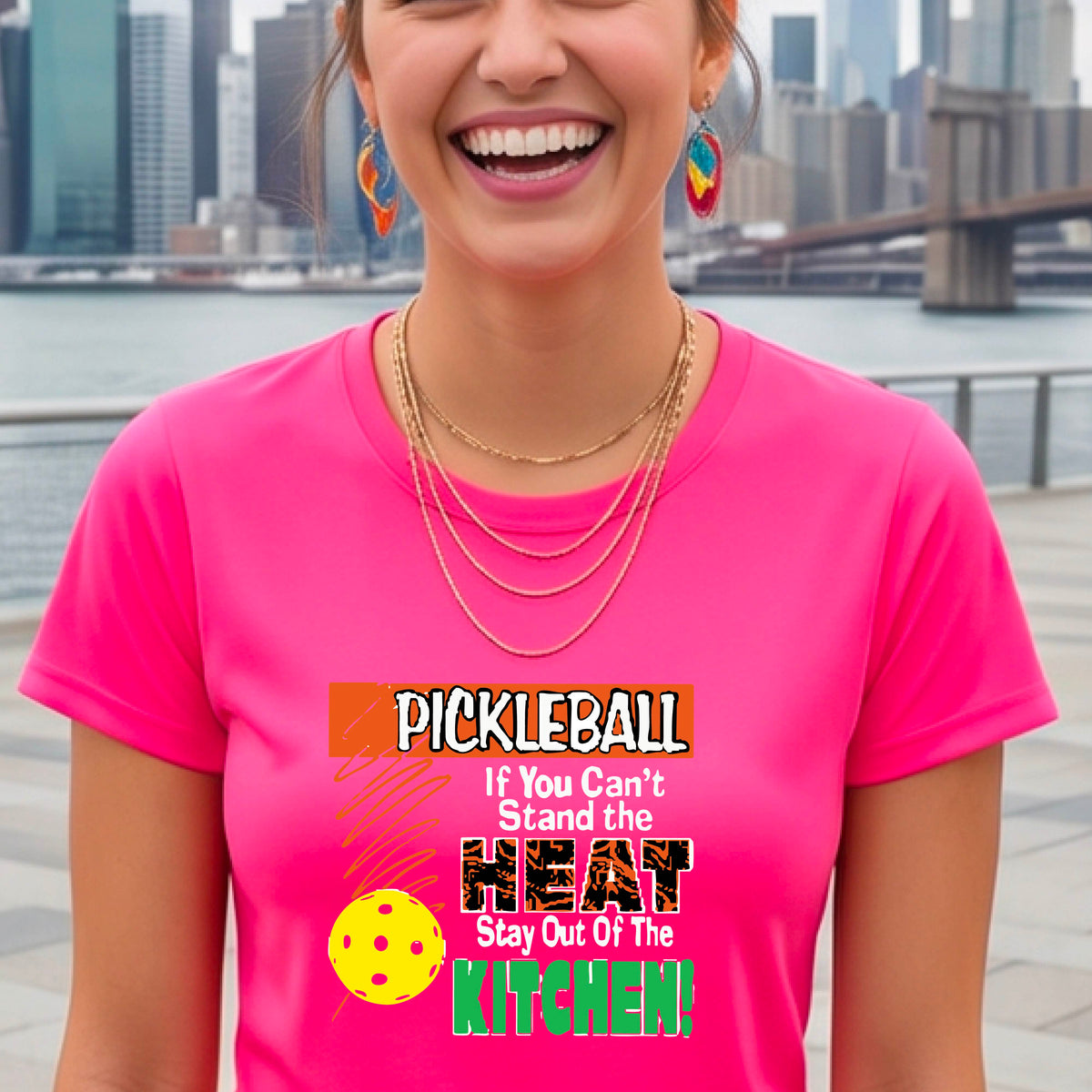 A woman wearing a pink short-sleeve t-shirt with a colorful graphic that reads 'PICKLEBALL If You Can't Stand the HEAT Stay Out Of The KITCHEN!' on a pickleball court background.