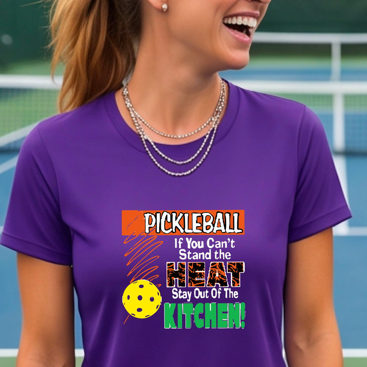 A woman wearing a purple short-sleeve t-shirt with a colorful graphic that reads 'PICKLEBALL If You Can't Stand the HEAT Stay Out Of The KITCHEN!' on a pickleball court background.