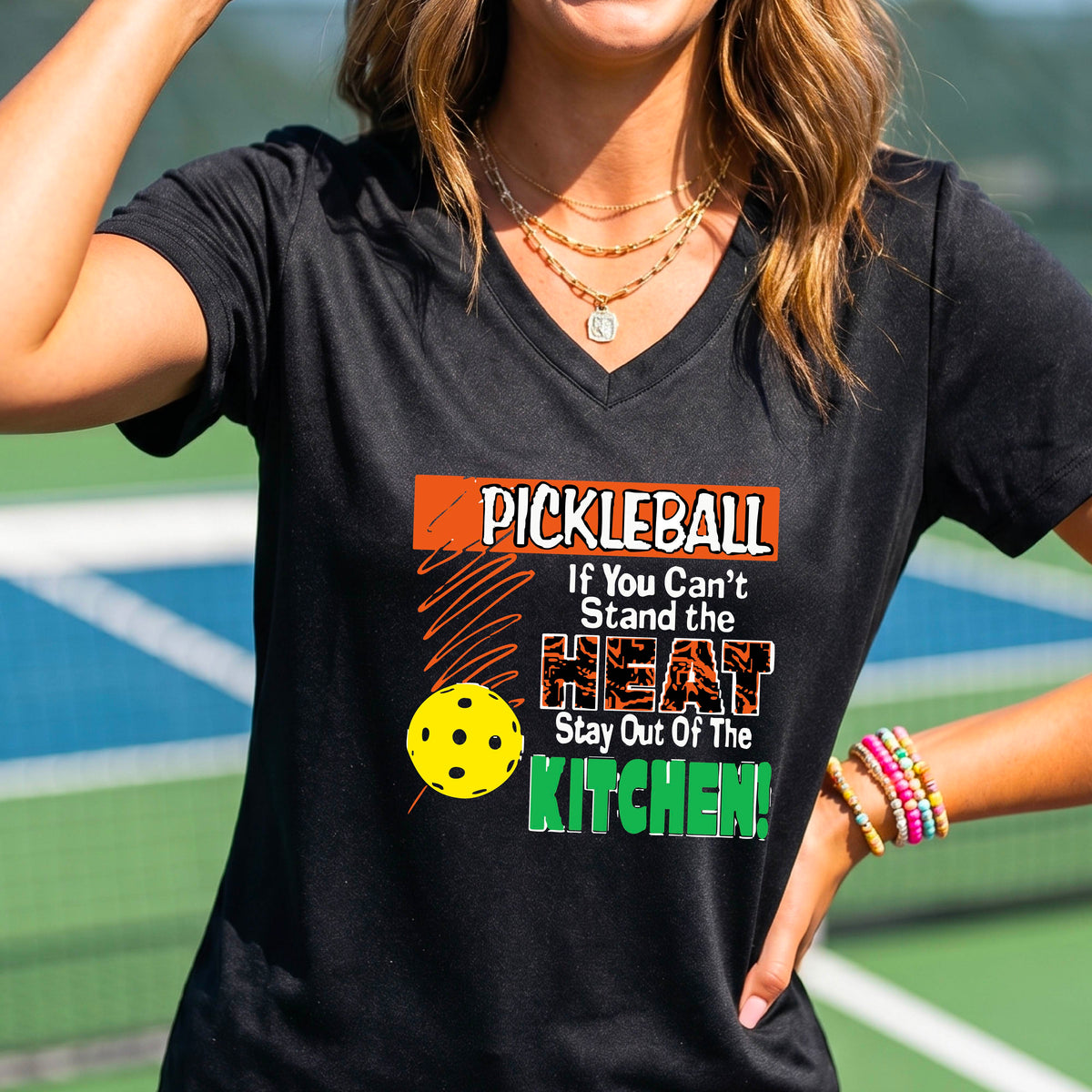 A woman wearing a black V-neck t-shirt with a colorful pickleball graphic and text that reads 'PICKLEBALL If You Can't Stand the HEAT Stay Out Of The KITCHEN!'. The background shows a blurred pickleball court.