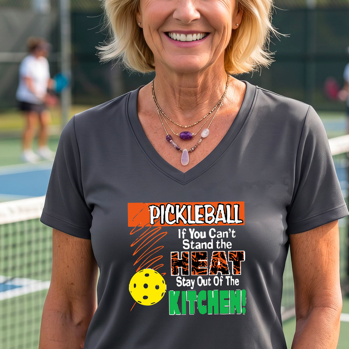 A woman wearing a gray V-neck t-shirt with a colorful pickleball graphic and text that reads 'PICKLEBALL If You Can't Stand the HEAT Stay Out Of The KITCHEN!'. The background shows a blurred pickleball court.