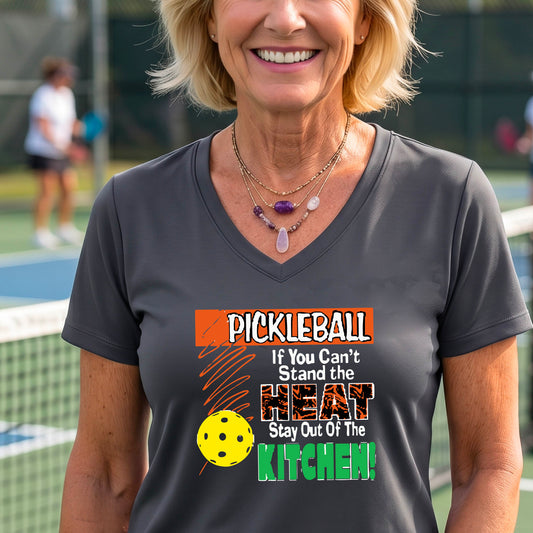 A woman wearing a gray V-neck t-shirt with a colorful pickleball graphic and text that reads 'PICKLEBALL If You Can't Stand the HEAT Stay Out Of The KITCHEN!'. The background shows a blurred pickleball court.