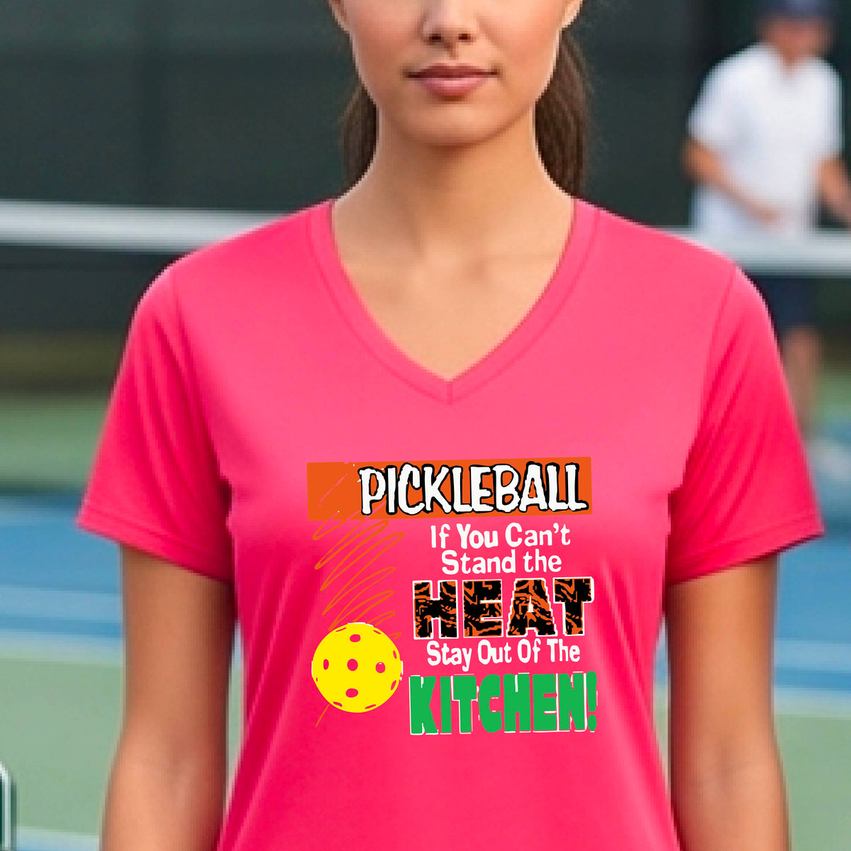 A woman wearing a bright pink V-neck t-shirt with a colorful pickleball graphic and text that reads 'PICKLEBALL If You Can't Stand the HEAT Stay Out Of The KITCHEN!'. The background shows a blurred pickleball court.