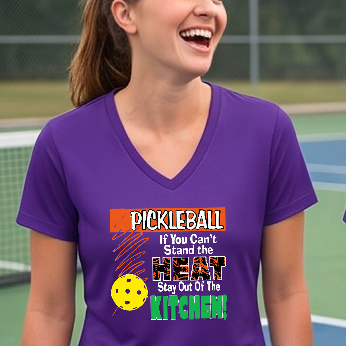 A woman wearing a purple V-neck t-shirt with a colorful pickleball graphic and text that reads 'PICKLEBALL If You Can't Stand the HEAT Stay Out Of The KITCHEN!'. The background shows a blurred pickleball court.