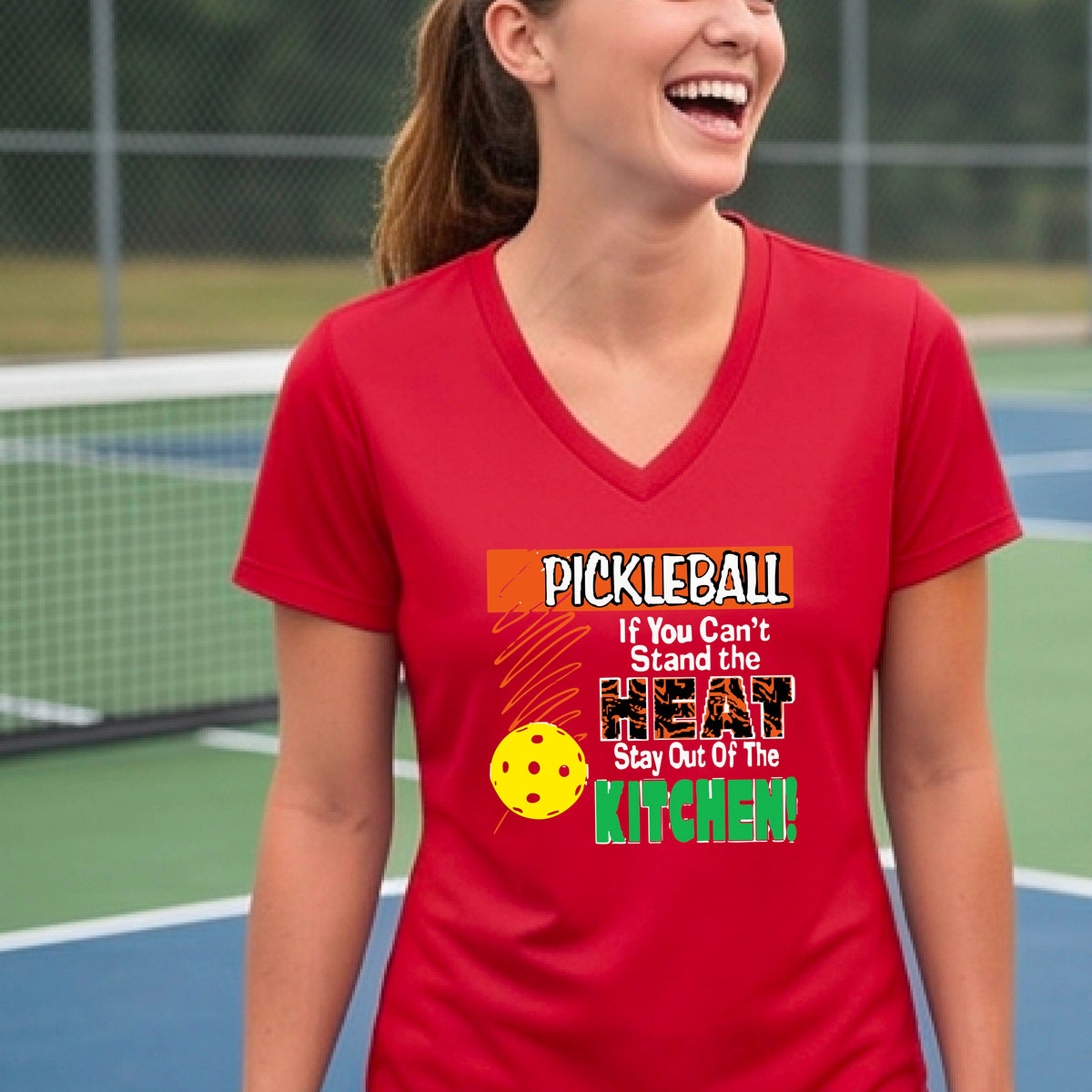 A woman wearing a bright red V-neck t-shirt with a colorful pickleball graphic and text that reads 'PICKLEBALL If You Can't Stand the HEAT Stay Out Of The KITCHEN!'. The background shows a blurred pickleball court.