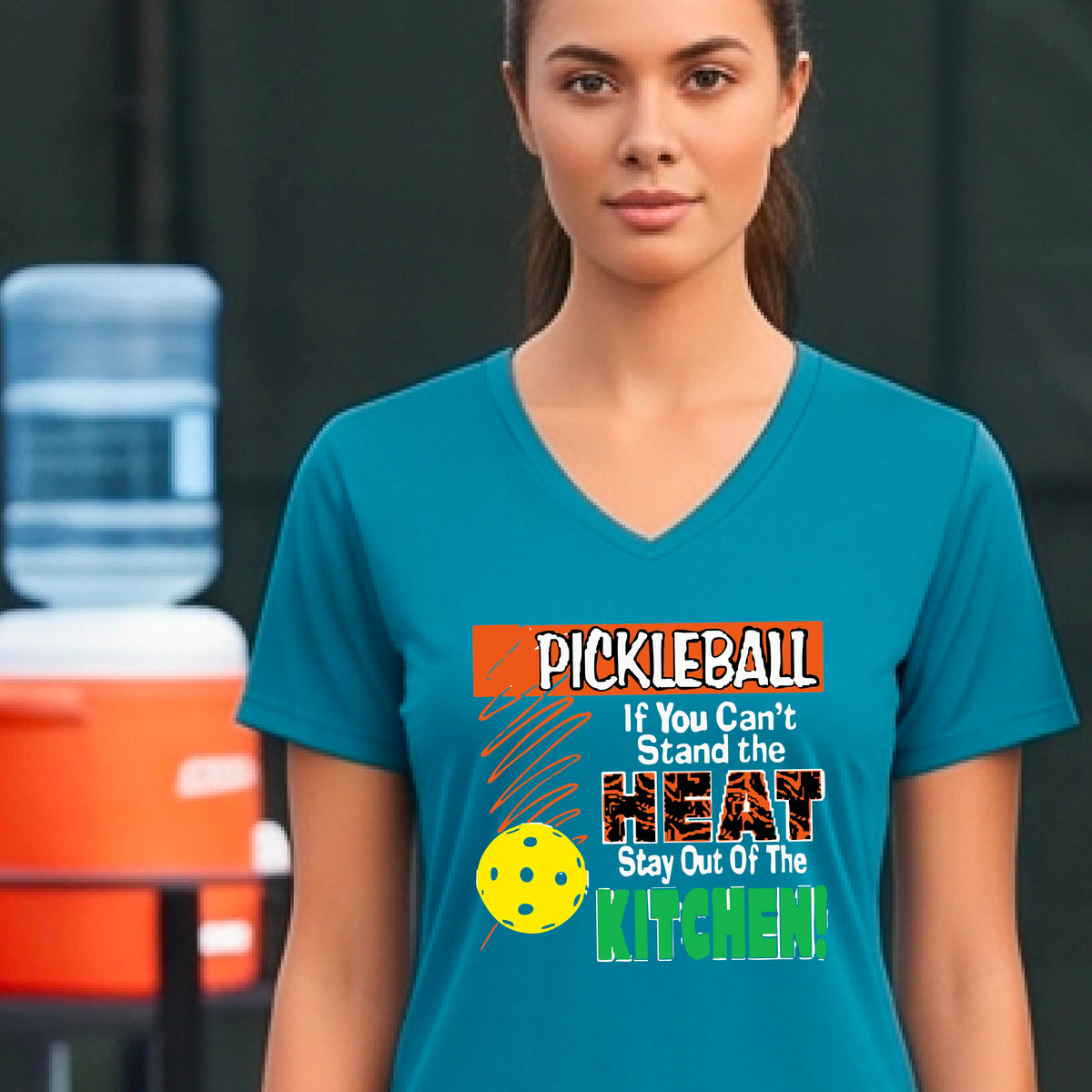 A woman wearing a teal V-neck t-shirt with a colorful pickleball graphic and text that reads 'PICKLEBALL If You Can't Stand the HEAT Stay Out Of The KITCHEN!'. The background shows a blurred pickleball court.