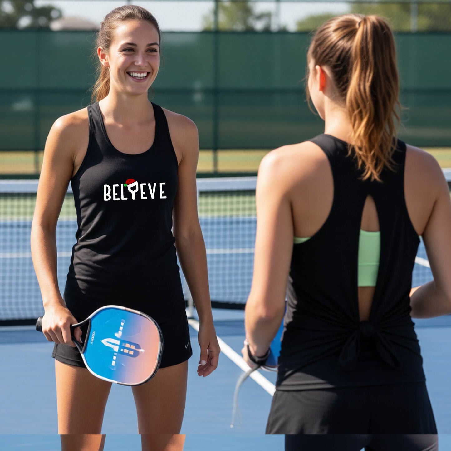 Two women on a blue pickleball court wearing matching black tank tops. One shows the front of the top with the word 'BELIEVE' and a small pickleball paddle Santa hat graphic, while the other shows the back with a tie detail.