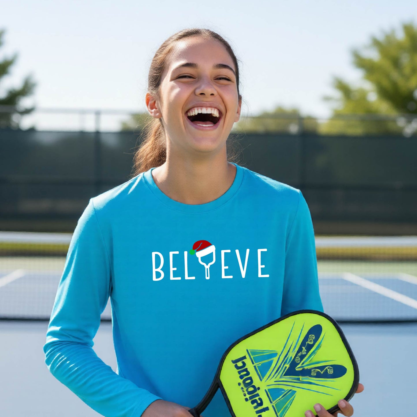 A young person laughing on a pickleball court wearing a bright blue long-sleeve shirt with 'BELIEVE' with i replaced with a pickleball paddle wearing a santa hat printed on the front.