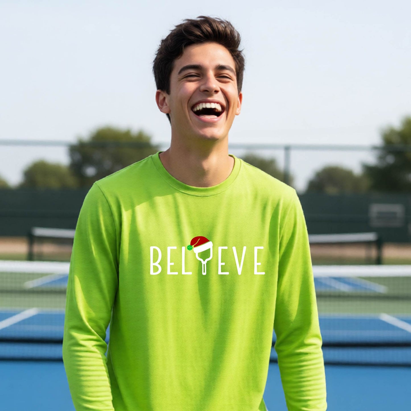 A young person laughing on a pickleball court wearing a bright lime green long-sleeve shirt with 'BELIEVE' with i replaced with a pickleball paddle wearing a santa hat printed on the front.