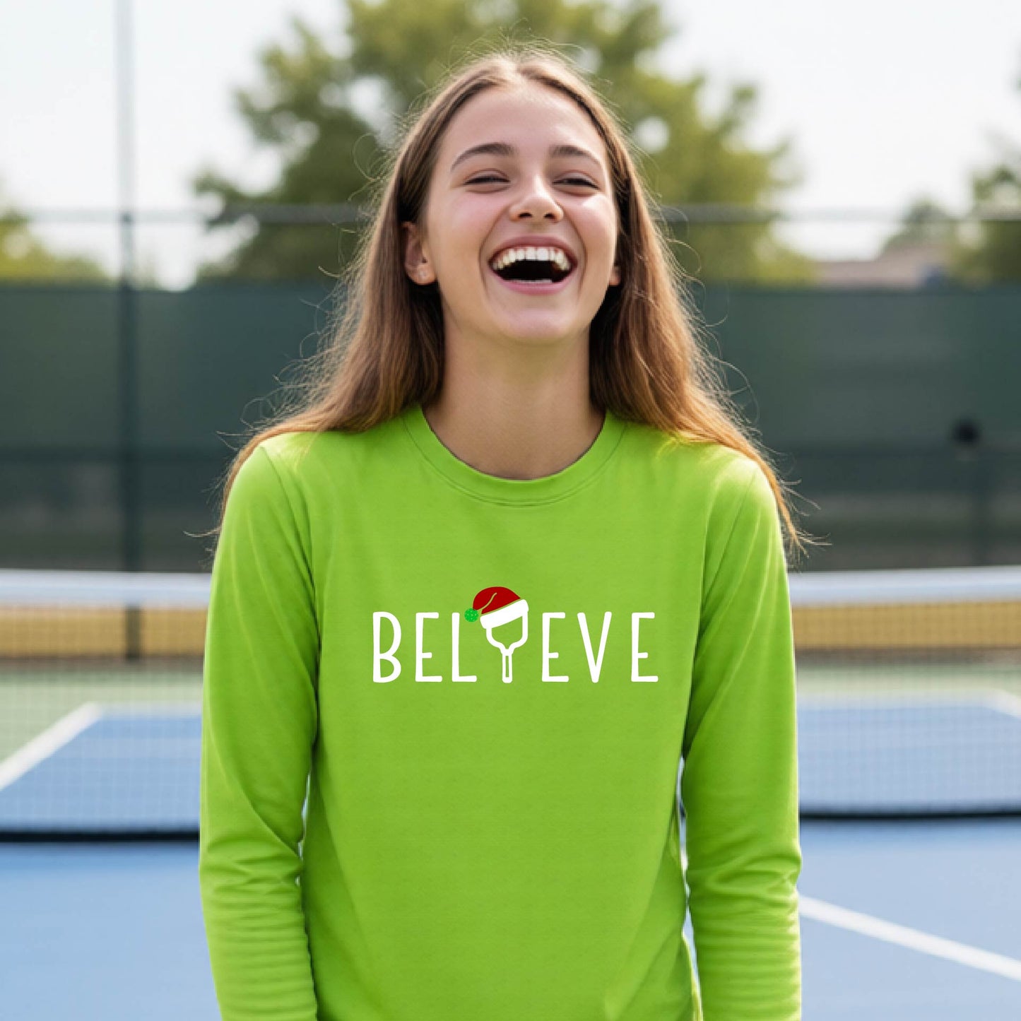 A young person laughing on a pickleball court wearing a bright lime green long-sleeve shirt with 'BELIEVE' with i replaced with a pickleball paddle wearing a santa hat printed on the front.